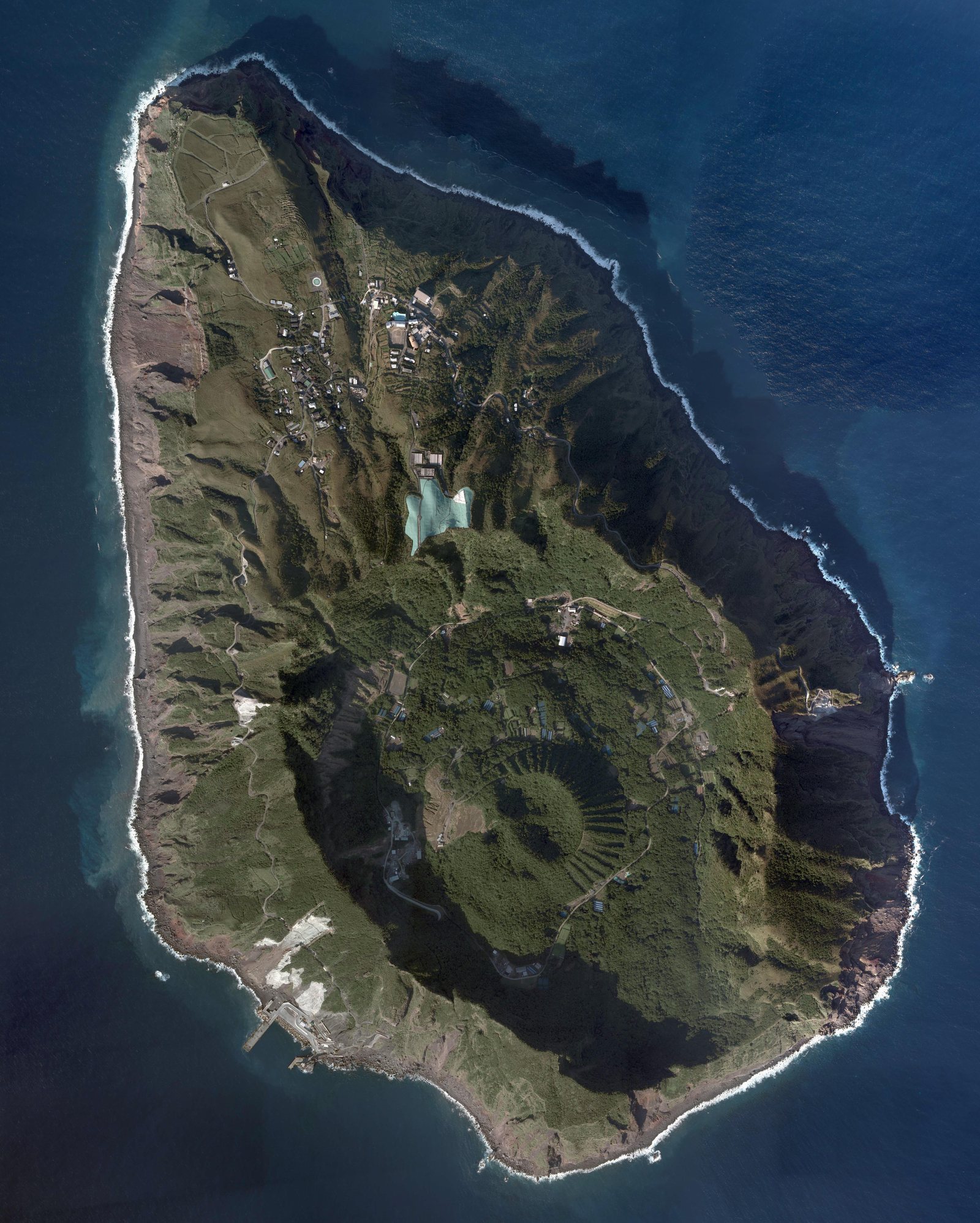 Vertical aerial view of Aogashima, an oval island ringed by sheer cliffs, its interior a green crater basin with a smaller forested cone standing inside it