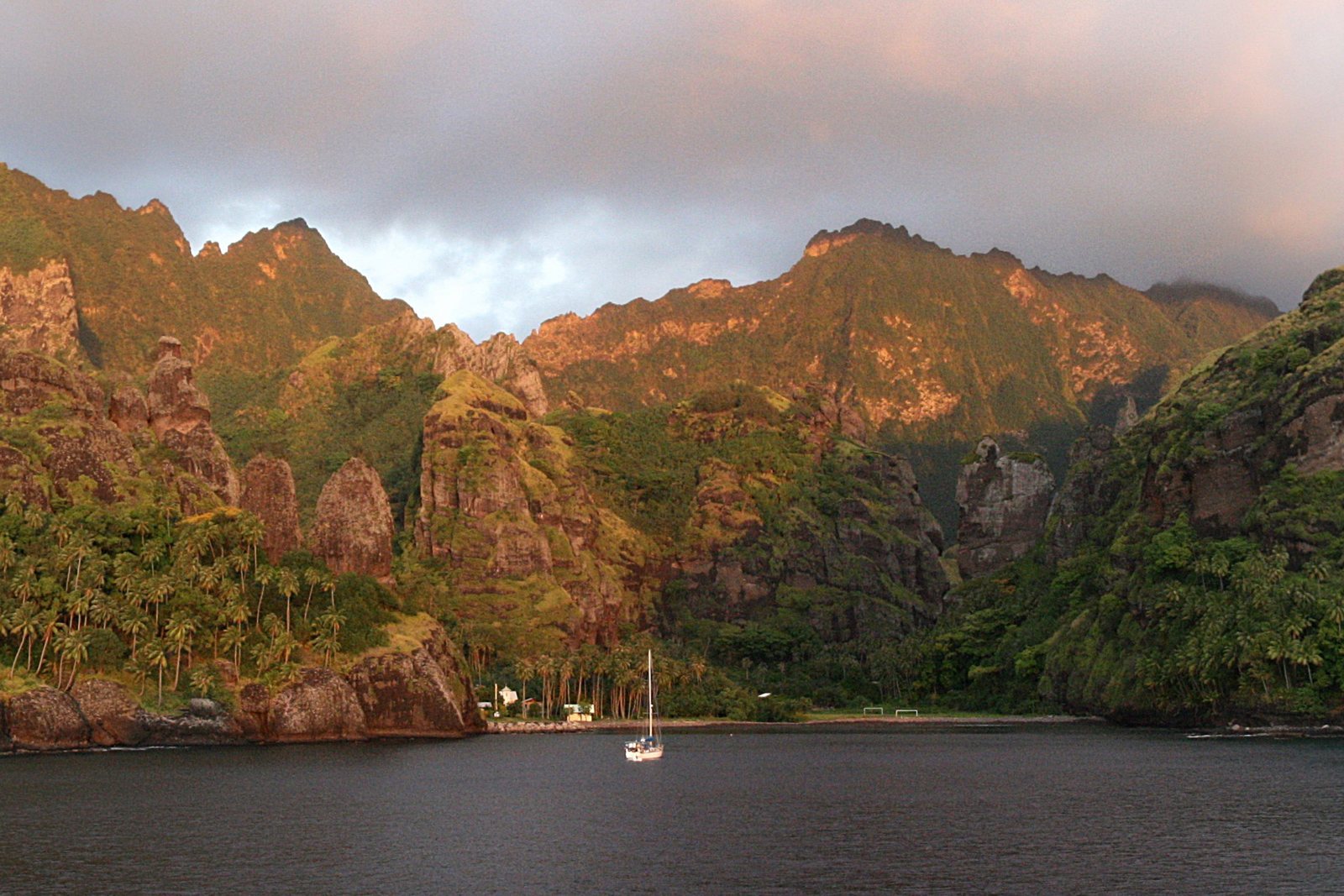 Eroded basalt spires catching late light above a palm-fringed shore, yachts anchored in the dark water of the bay below
