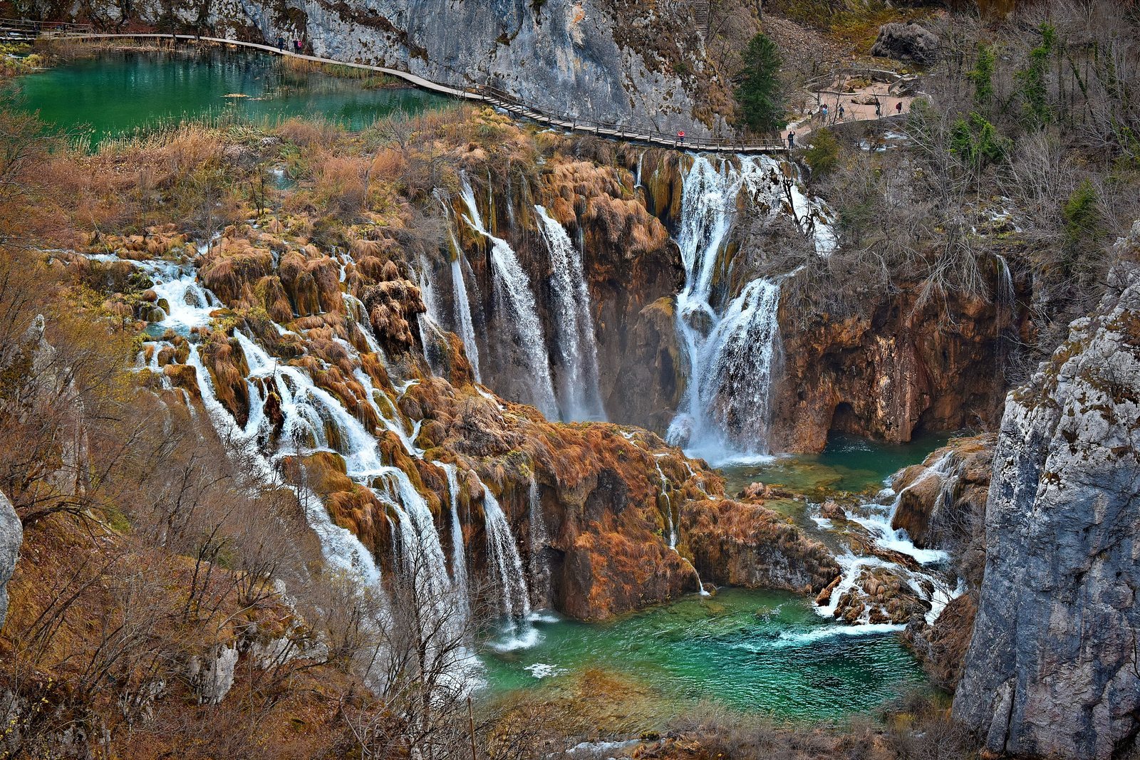 A row of waterfalls spilling over a rock barrier from a green upper lake into a turquoise pool below, a wooden boardwalk along the rim