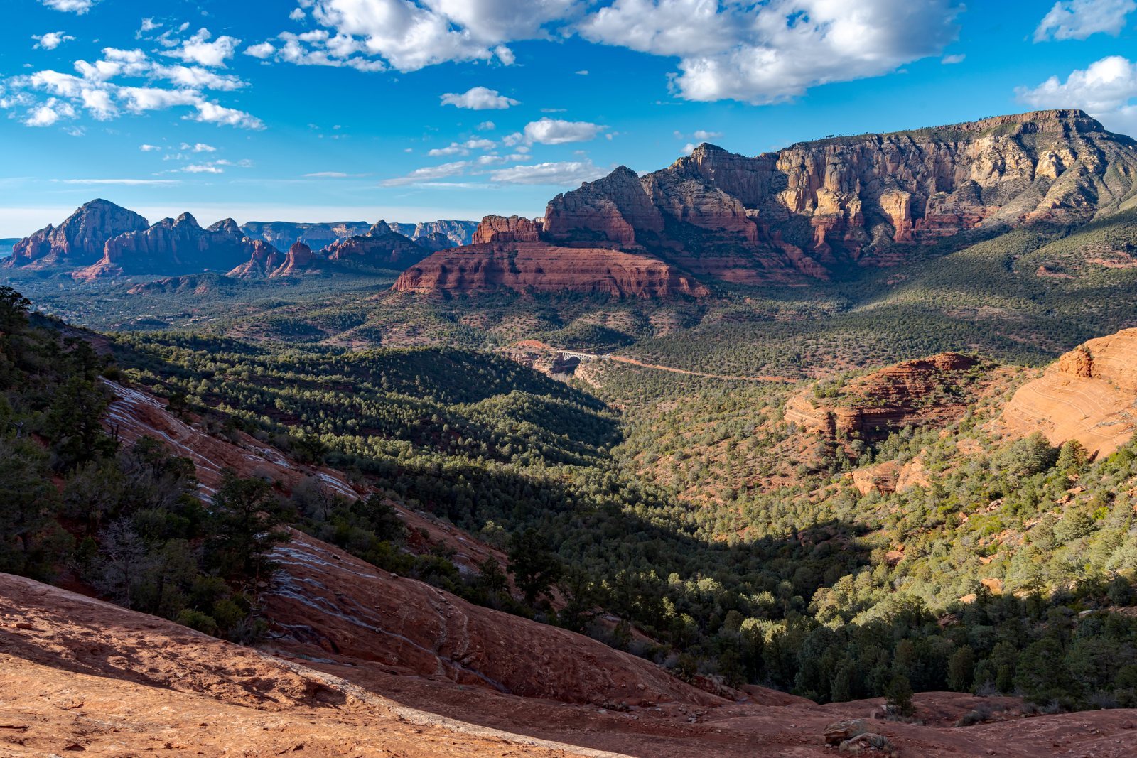 Layered sandstone buttes and cliffs stained deep red standing above a wide green valley of juniper and pine under a blue sky