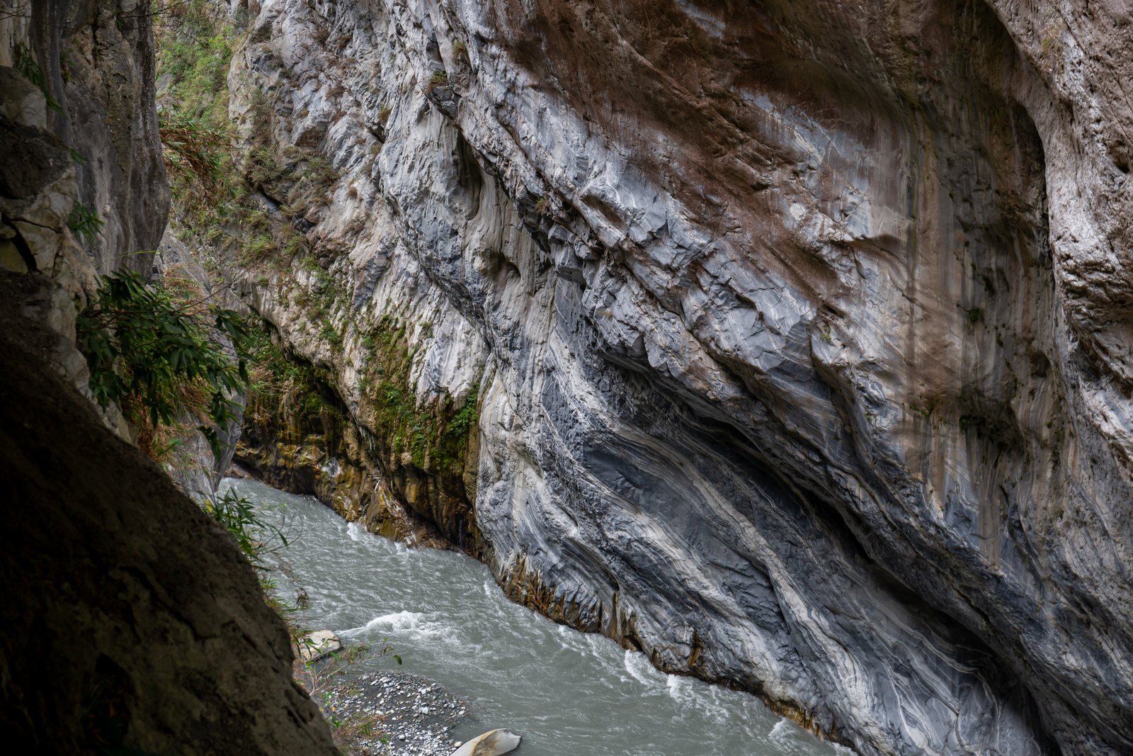 A wall of grey and white banded marble, its layers folded and swirling, dropping straight into pale green river water at the foot of the cliff